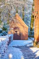 Exterior of an old church building with snow covered trees and pathway. Small chapel in snowy landscape with sun shining on brick wall. Countryside with European style architecture in winter scene