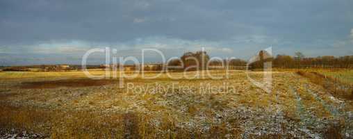 Landscape of a field on a winter day with a bit of snow on the grass and a cloudy sky. Dry tan grass in an open meadow. Rural countryside with brown pasture melting snow and trees on horizon