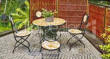 Set of outdoor dining furniture. Mosaic tile and iron chairs and table on a grey brick patio in a lush garden. Cozy enclosed relaxation area outside. Private location to enjoy the warm summer weather