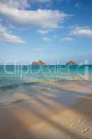 A landscape view of crystal clear ocean water with sand and island. A bright sky with white clouds. A picture with shadows and footsteps on the sand. A shot of wavy blue ocean displaying mix texture.