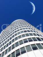 Half a moon over a building skyscraper, shining in bright blue sky. Below the crescent of quarter moon over a tall modern structural architecture, foreboding doomed capitalism or an economical crisis