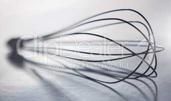Blurred closeup of stainless steel balloon whisk on a grey table in a modern kitchen. Metallic, eleven wire, egg beater ready to cook against a white background. A cooking utensil perfect for mixing