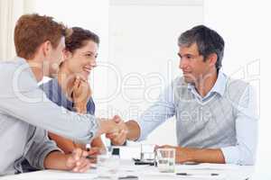 This merger will benefit both companies. two businessmen shaking hands while in a meeting with a female coworker.