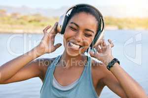 Excited woman smiling and listening to music with water in the background. Happy young female enjoying tunes on her online streaming playlist while outdoors. Using wireless headphones play songs