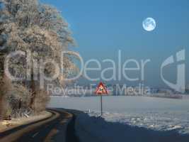 The winding road on a rural winter landscape with a sign on the side of the empty road for safety. A slippery and wet road surrounded by snow on a cold winter evening near a forest with ice frost