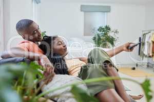 Young african american couple changing channels on remote and watching television together on sofa at home. Girlfriend relaxing on boyfriends lap while enjoying entertainment shows, series and movies