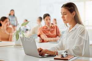 One young hispanic business woman working on a laptop in a busy office with her colleagues in the background. Focused entrepreneur browsing the internet while planning ideas at her desk in a creative startup agency