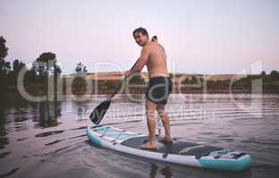 Young active caucasian man smiling and using his stand up paddle board on a lake out in nature. Young male enjoying a summer day at dawn on a paddle board. Life is better when youre paddling in the water