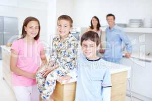 Getting ready to eat a wholesome breakfast. Three children in the kitchen with their parents standing blurred in the background.
