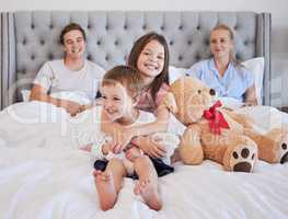 Adorable little girl embracing her younger brother with her teddybear next to her while sitting on a bed with per parents in the background. Loving young caucasian family bonding and spending time together in the morning. Having a big sister means youll a