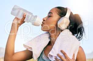 An active fit woman wearing wireless headphones drinking water from a bottle after exercising outdoors. Female athlete quenching thirst and cooling down with a towel after training workout outside