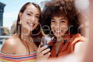 Two beautiful mix race women smiling taking a selfie while enjoying a glass of red wine while outdoors at a party. Hispanic female with cool afro hairstyle enjoying her time with a friend on a sunny day while enjoying a beverage