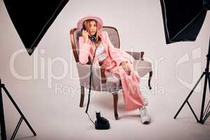 Stylish young mixed race female posing in trendy pink clothes sitting on a chair and making a call with a retro telephone for a studio photoshoot. Female model on the set of an editorial fashion shoot