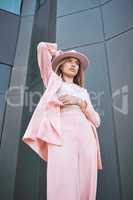 Fashionable young hispanic model wearing a stylish trendy pink suit and hat while posing outside against a modern building in the city