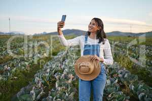 Woman farmer taking a selfie on her smartphone while standing in a cabbage field. Young brunette female with a straw hat using her mobile device on an organic vegetable farm