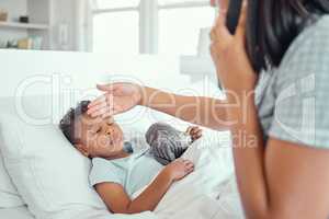 Sick little boy in bed while his mother talks on the phone calling doctor for help to bring fever down. Young single parent feeling sons forehead to check temperature. Mixed race child feeling ill and lying in bed while his mother checks fever