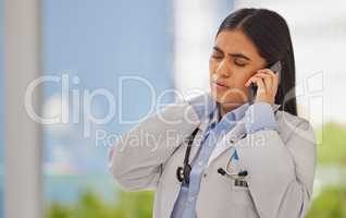 Stressed doctor talking on cellphone in a clinic. Indian healthcare professional standing alone and suffering from neck pain on a busy shift. Medical specialist booking appointment for physiotherapy