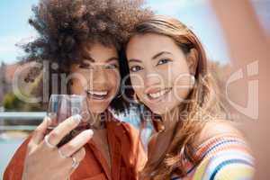 Two beautiful young mixed race women standing together and taking a selfie while bonding outside. African american with an afro drinking wine with her asian friend. Capturing memories for social media