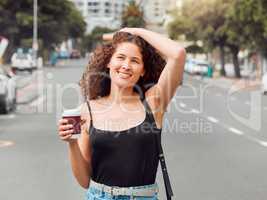 One happy beautiful young mixed race woman exploring the city while holding a takeaway coffee and touching her curly brunette hair. Hispanic travelling woman enjoying the view downtown on the weekend
