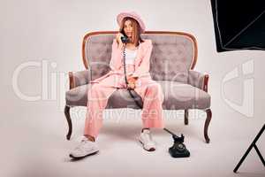 Young mixed race female posing in trendy clothing while sitting on a couch and making a call with a landline phone against a grey studio background. Confident hispanic woman posing in pink clothes