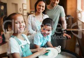 Young happy mixed race parents washing the dishes with their son and daughter in the kitchen at home. Little brother and sister helping their mom and dad clean the dishes. Girl washing a plate