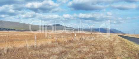 Harvested farm land beside a highway on cloudy day. Empty wheat field against a blue sky horizon. Rural agriculture with dry pasture near mountains. Grass growing beside an empty road in the country