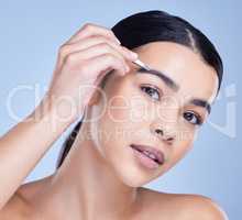 Studio portrait of a beautiful mixed race young woman with glowing skin posing against blue copyspace background while tweezing her eyebrows. Hispanic model using a tweezer for hair removal