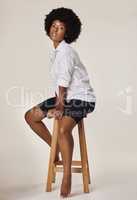 Studio portrait of a young stunning African American woman with a beautiful afro.Confident black female model showing her smooth complexion while sitting on a chair and posing against a grey background