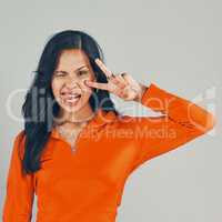 Portrait of mixed race woman isolated against grey studio background with copyspace. Young playful hispanic standing alone, making peace sign and symbol with hand gesture and winking with tongue out