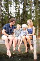 Sharing precious family moments. Cute young family laughing together while sitting on a lakeside jetty - outdoors.