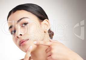 Beautiful young woman popping a pimple standing against a grey studio background alone. One female touching her face while standing against a background