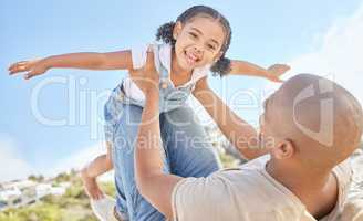 Father and daughter bonding together outdoors. Portrait of adorable little girl from below having fun pretending to fly like a superhero with arms outstretched while being held by her loving dad.