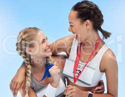 Two cheerful athletic women embracing while holding gold medals from competing in sports event. Joyful fit active athletes feeling proud after winning in running race. Track runners achieving goal