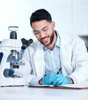 One handsome young mixed race man wearing gloves and a labcoat and looking at medical samples on a microscope in a lab. A male scientist wearing safety goggles and smiling while writing in a notebook