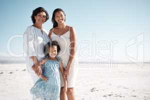 Smiling mixed race family standing together on a beach with copyspace. Happy hispanic grandmother bonding with granddaughter over weekend. Adorable little girl enjoying free time with single mother