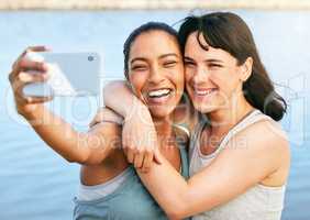Two female friends taking selfies after a workout in nature and standing in front of a lake using a smartphone. Young Latino female using her phone to take a selfie of herself and her friend