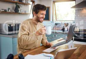 Happy young caucasian man drinking coffee while working on laptop in kitchen, checking his email or searching information while doing freelance work at home. Smiling young male using internet banking service