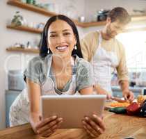 Portrait of beautiful young woman browsing on digital tablet while her boyfriend cooks in the background. Young hispanic female searching for recipe to follow online while cooking at home with her husband