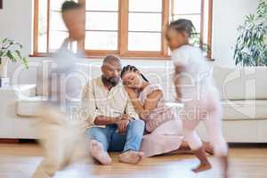 Tired African American couple sitting on the lounge floor looking tired and drained while their energetic children plays around them. Two black siblings having fun while their exhausted parents relax