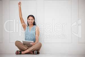 Cheerful young mixed race woman browsing on a digital tablet device and cheering while sitting on the floor in an office. One successful female celebrating an awesome achievement with her fist raised