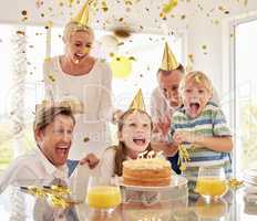 Cheerful generational happy family wearing party hats while celebrating little girls birthday with confetti and cake at home. Excited kids, parents and grandparent enjoying special surprise event