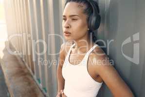 One fit young hispanic woman wearing headphones and taking a rest break to catch her breath after a run or jog in an urban setting outdoors. Female athlete looking tired after intense cardio exercise