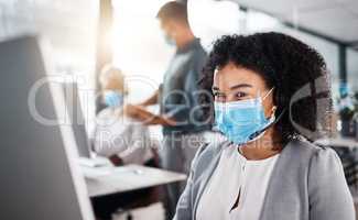 Mixed race call centre telemarketing agent wearing face mask as safety protocol and talking on a headset while using computer in an office. Female consultant operating helpdesk for customer service and sales support during covid pandemic