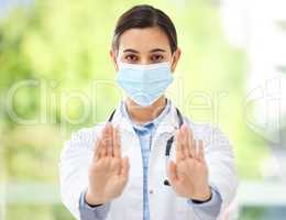 Portrait of a hispanic female doctor wearing a mask and showing a stop gesture with her hand while wearing a stethoscope in a hospital office.