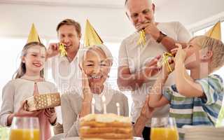 Senior woman celebrating her birthday with family at home, wearing party hats and blowing whistles. Grandma looking at birthday cake and looking joyful while surrounded by her grandkids and and son