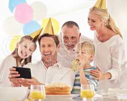 A happy family celebrating a birthday with a party, wearing hats and taking a selfie using a phone. Mature man taking a photo of his father, wife and children at a party while making special memories