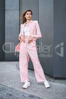 Full body young beautiful mixed race woman standing alone in a city and posing in fashionable clothes. Serious hispanic in trendy pink suit showing street style. Feeling bold, confident with fashion