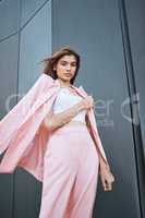 Below view portrait of young beautiful mixed race woman standing alone in a city and posing in fashionable clothes. Serious hispanic in trendy pink suit showing street style. Feeling bold, confident