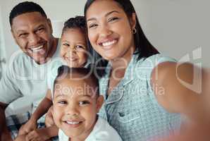 Photo of a loving mother holding phone and taking selfie or recording funny video with her family at home. Happy mixed race family with two children and parents posing together for a family picture on a mobile phone