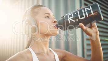 One fit young hispanic woman wearing headphones and taking a rest break to drink water from a bottle while exercising outdoors. Female athlete quenching thirst and cooling down after run and training workout in an urban setting